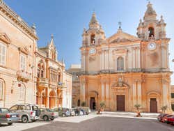 A beige-coloured cathedral under a blue sky with cars parked in front of it