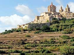A city at the top of a hill surrounded by grass under a blue sky with white fluffy clouds
