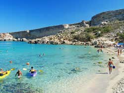 A sandy beach full of people kayaking and swimming in the water under a blue sky