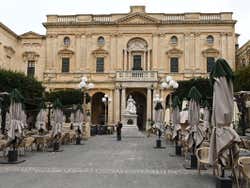 A square with a statue in front of a large building and several outdoor seating areas